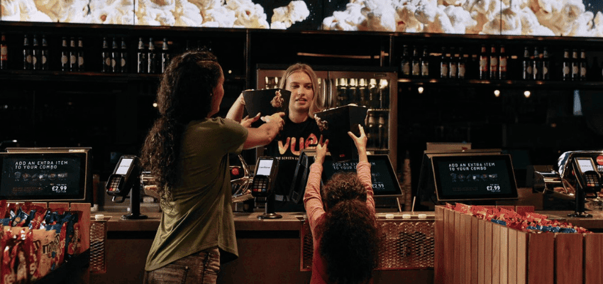 A child reaching for popcorn at the Vue cinema counter while her parent is served by a friendly staff member, highlighting inclusive family cinema experiences and affordable Vue Offers for Disabled People.