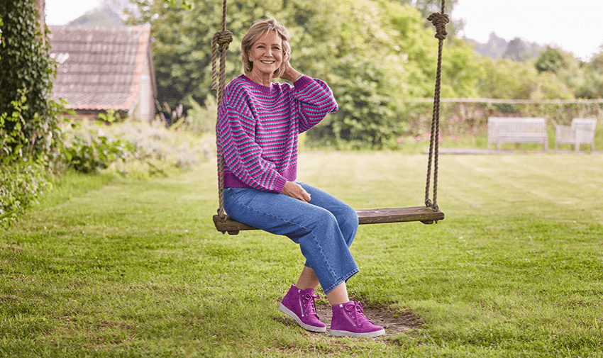 A woman smiling and sitting on a wooden swing in a garden, wearing a purple jumper, blue jeans and purple trainers