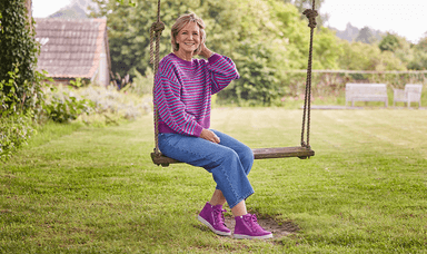 A woman smiling and sitting on a wooden swing in a garden, wearing a purple jumper, blue jeans and purple trainers