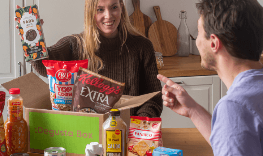 A man and woman smile and point at each other excitedly in a kitchen while unpacking a Degusta Box containing various food items such as cereal, snacks, and sauces, arranged on a counter. The box is filled with items like pasta, popcorn, and cooking ingredients.