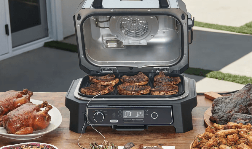 Ninja Woodfire electric BBQ grill packed with sizzling steaks, surrounded by roast chicken, charred vegetables and crispy sides, showing off outdoor cooking ease with mobility discounts and offers.