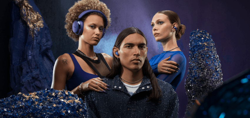 A striking image featuring three people surrounded by large, dark blue and gold crystalline formations. The person in the center wears a dark jacket and blue earbuds. The person on the left wears a blue top and blue over-ear headphones. The person on the right wears a sheer blue top and blue/purple earbuds.