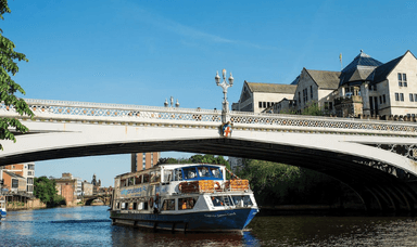 A sightseeing boat cruises along a river under an ornate bridge on a clear day. In the background, there are historic buildings and lush trees lining the riverbank. People can be seen walking across the bridge.