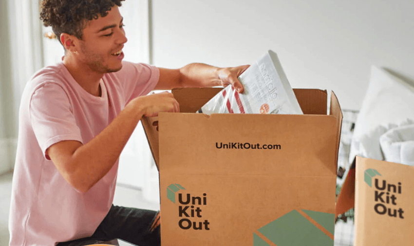 Young man in a light pink T-shirt smiling while unpacking a Uni Kit Out student starter kit on a bed, with household essentials such as a mixing bowl, cheese grater, and folded grey towels in the foreground, highlighting moving essentials for disabled students and Uni Kit Out offers in the UK.