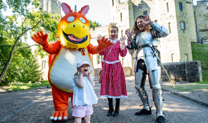 Smiling young girl posing with characters at Warwick Castle, including a large orange Zog dragon mascot, a woman dressed as a princess, and a man in knight’s armour, promoting accessible family days out and UK attraction discounts for disabled people.