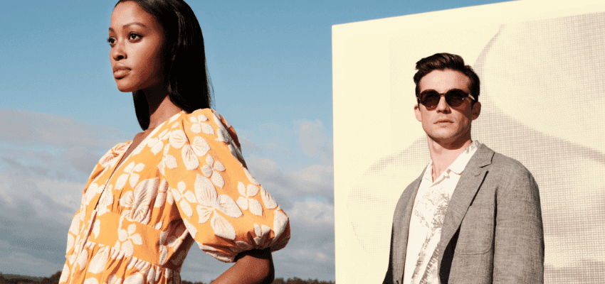 A woman in an orange floral dress and a man in a gray blazer with sunglasses stand outdoors against a sky backdrop and a large white screen. The woman faces forward, while the man looks slightly to the side. Both individuals appear confident and stylish.