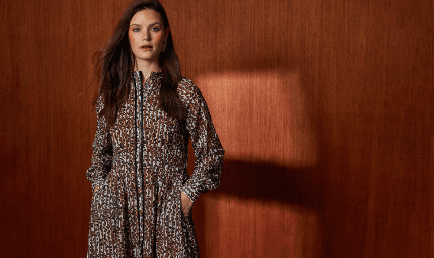 A woman stands against a wooden wall. She is wearing a long-sleeved, leopard print dress with her hands in the pockets, looking confidently at the camera. The lighting casts a warm tone across the scene.