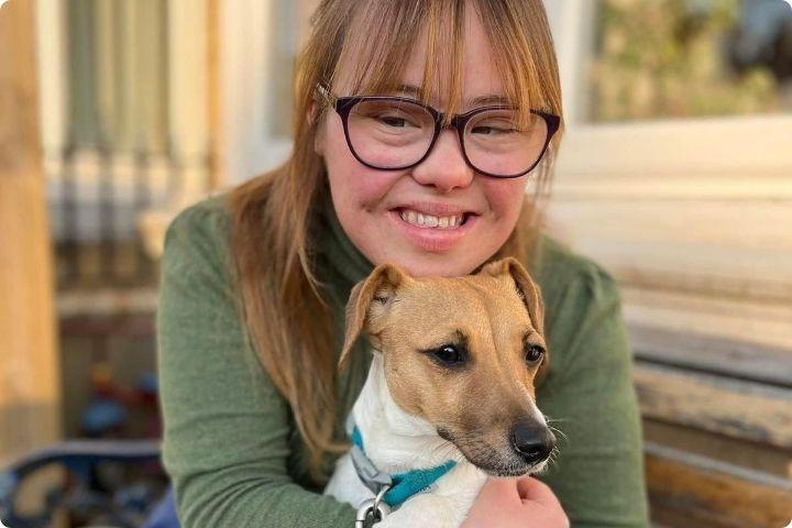 Florence Halliwell is a UK based land artist with Down's syndrome, she has dark blonde long hair with a fringe and wears glasses, she has a sage green long sleeved top on and is smiling and hugging a small terrier dog who has a blue colour on