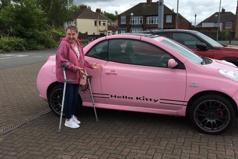 Sue, a disabled woman living with cancer, standing with crutches beside her pink car.
