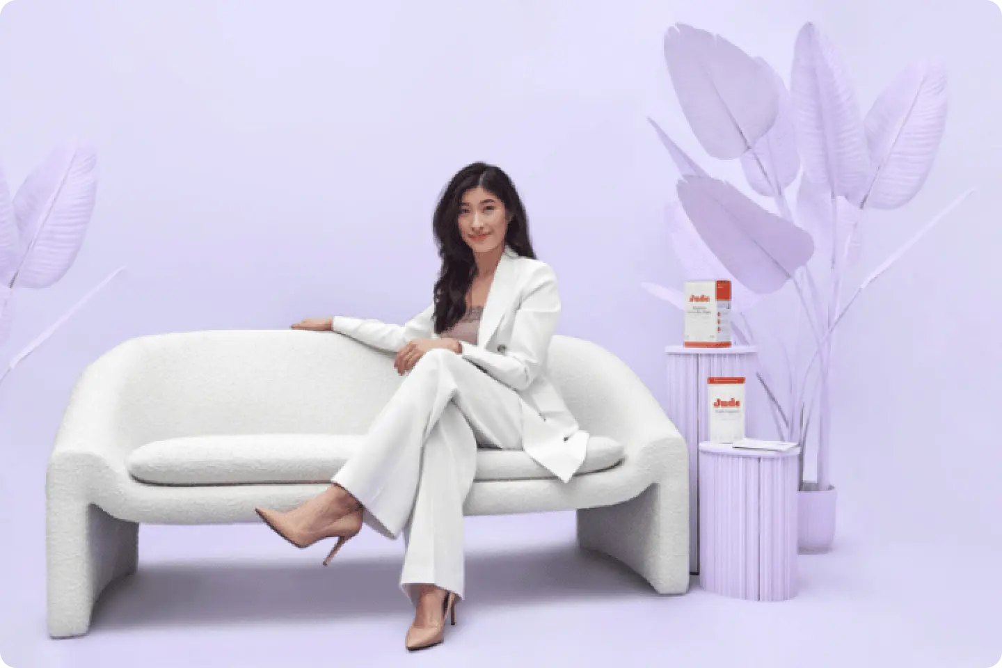 A woman in a white suit sits on a modern, white couch in a room with light purple walls and decor, including large, artificial leaves and two cylindri