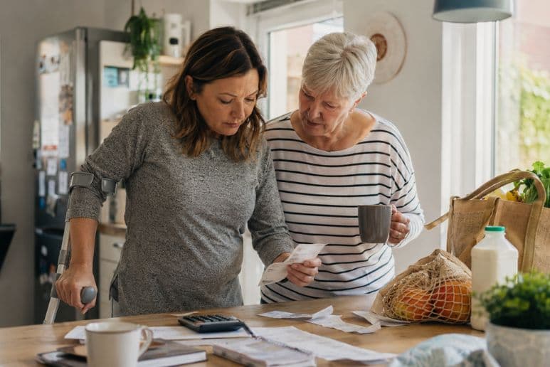 Disabled woman using a crutch reviewing grocery receipts and household costs with support from another woman in a kitchen.