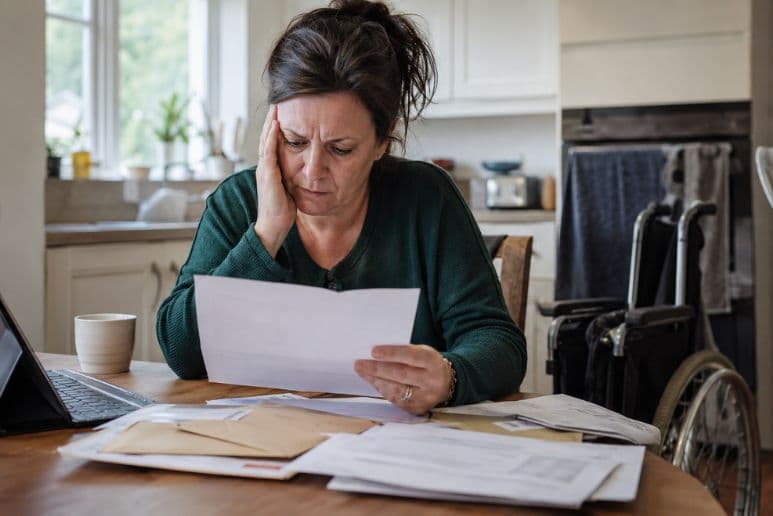 Worried disabled woman reading a DWP benefit letter at home with paperwork, laptop and mobility aids nearby.