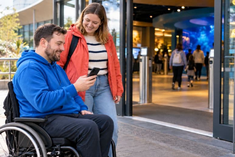 Wheelchair user checking a Digital Access Pass on their phone outside an accessible UK attraction with a companion.