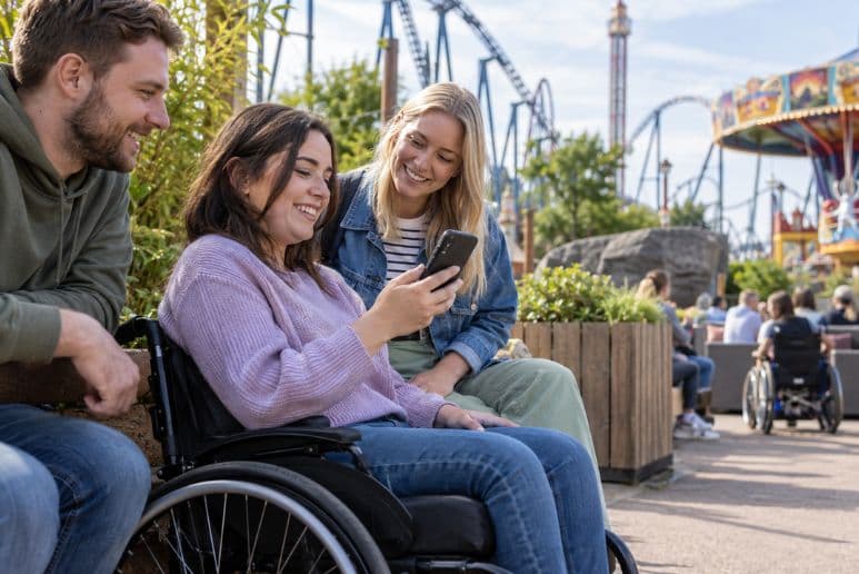 Disabled theme park visitor using a smartphone with friends while waiting away from the main ride queue.
