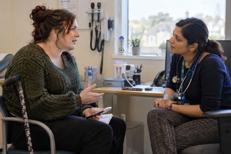 Disabled young woman with a walking stick discussing PIP medical evidence with a GP in a UK surgery.