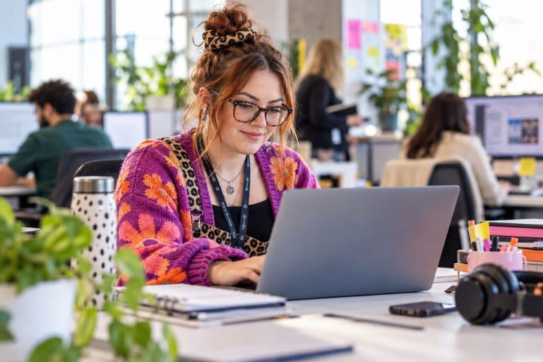 Neurodiverse woman working on a laptop in a modern office with colleagues in the background.